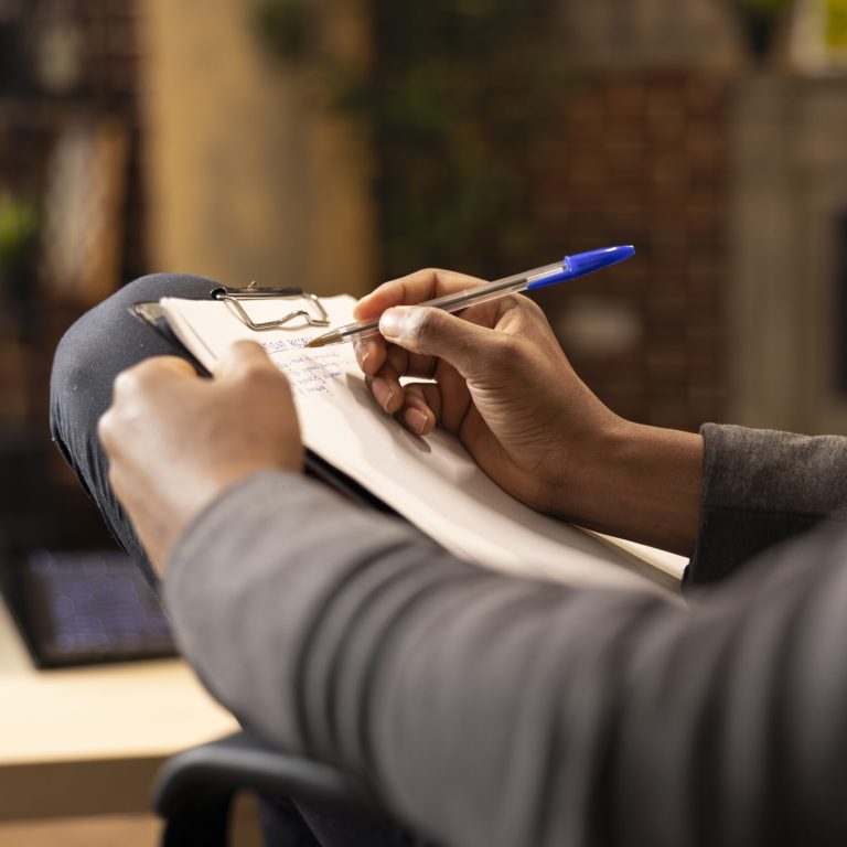 Black male psychologist writing in notepad, taking notes at private clinic. African american therapist with pen in hand, documenting on clipboard during session in brick wall office.